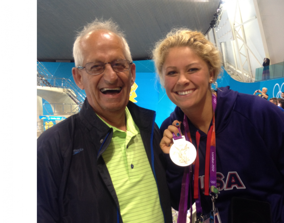 The late Ted Beisel next to Liz Beisel. They are in an indoor swimming pool stadium. Liz is holding her Olympic medal. Both are smiling.