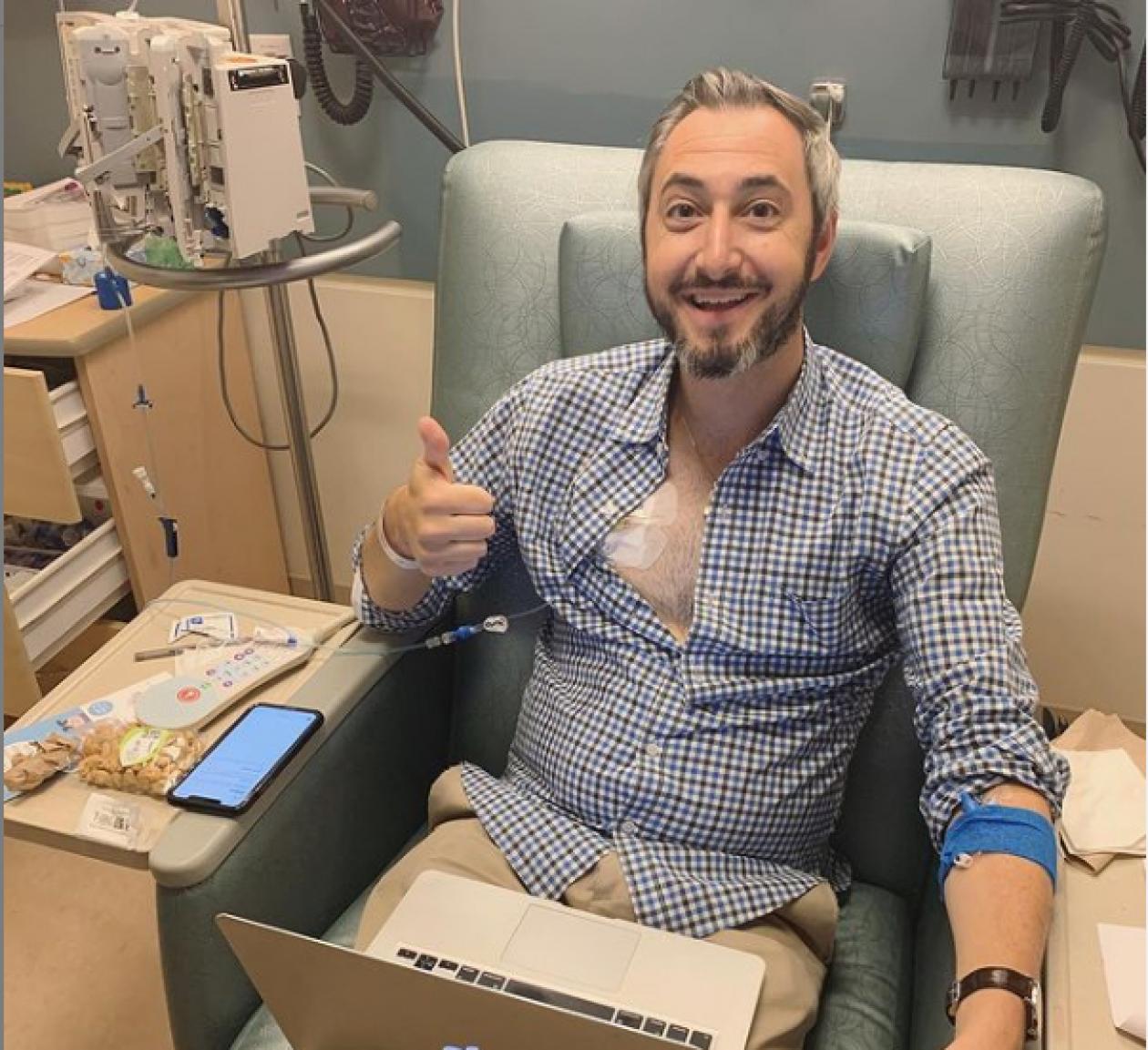 Dave Thau in a clinical setting, smiling and holding a thumbs-up. He has a monitor attached to his chest and is sitting, with a laptop.