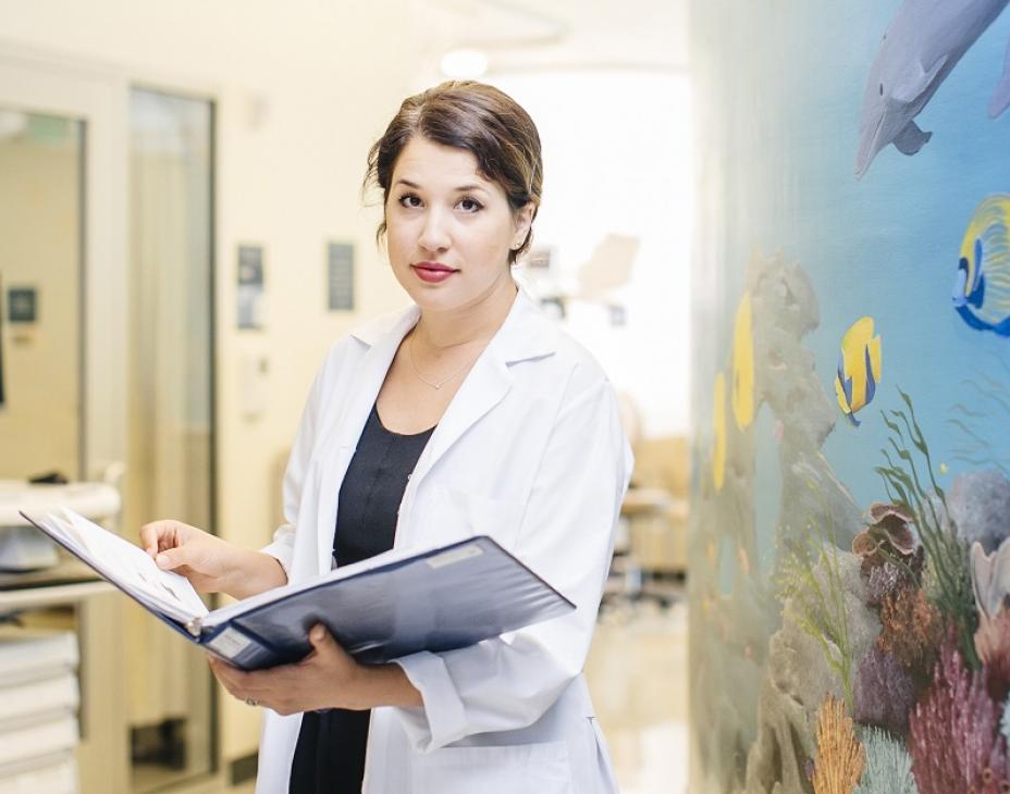 Dr. Elysia Alvarez in a clinical setting, holding an open book, while looking at the camera. The wall to her left is an aquarium painting with marine life. The background features a hospital hallway. She is wearing a white coat.