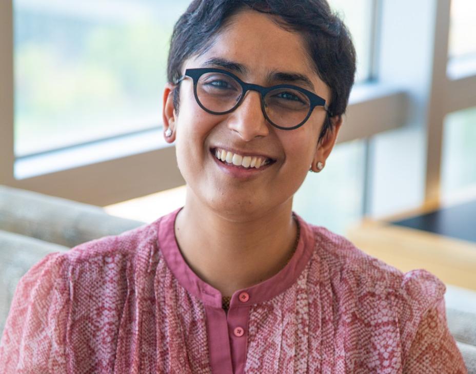 Dr. Salvia Jain smiling facing forward. She's sitting in a bright room with natural light. She's wearing a light pinkish-red shirt and reading glasses, and has short black hair.