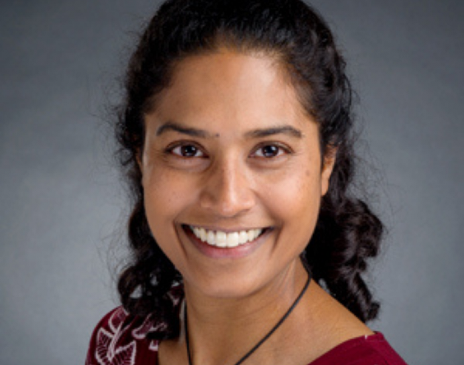 Dr. Sheena Mukkada facing forward and smiling. She has shoulder-length black hair and is wearing a dark-red shirt with a black necklace, against a plain gray background.