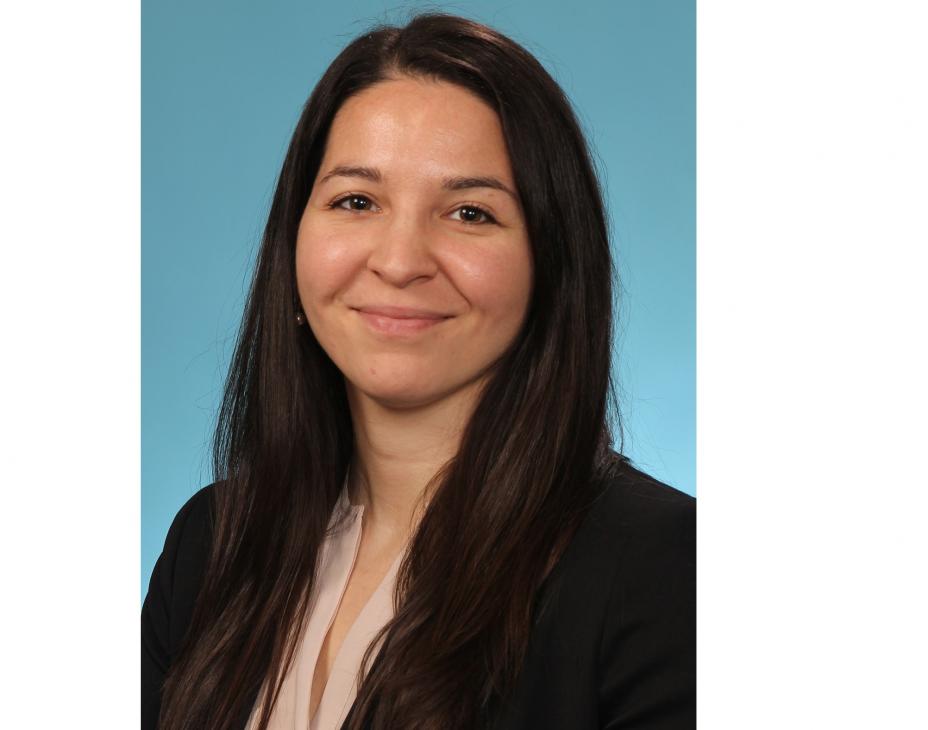 Headshot of Dr. Stephanie Markovina. She has long dark-brown hair and is wearing a black blazer with tan blouse, against a light-blue background. She is smiling facing forward.