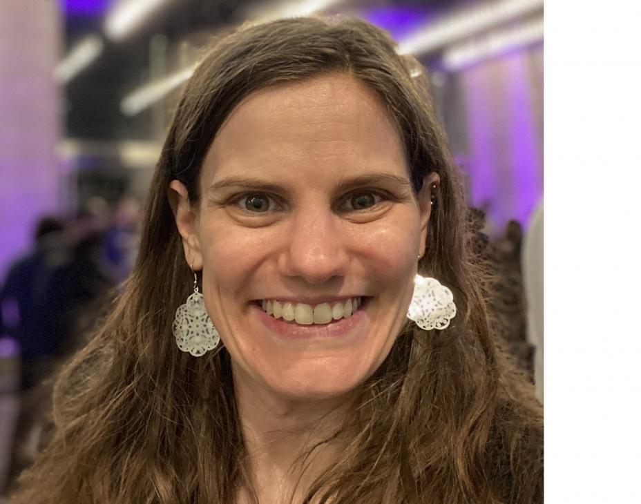 Carly Flumer selfie, smiling facing forward outside of a concert venue. She has long brown hair and is wearing silver earrings and a black jacket.