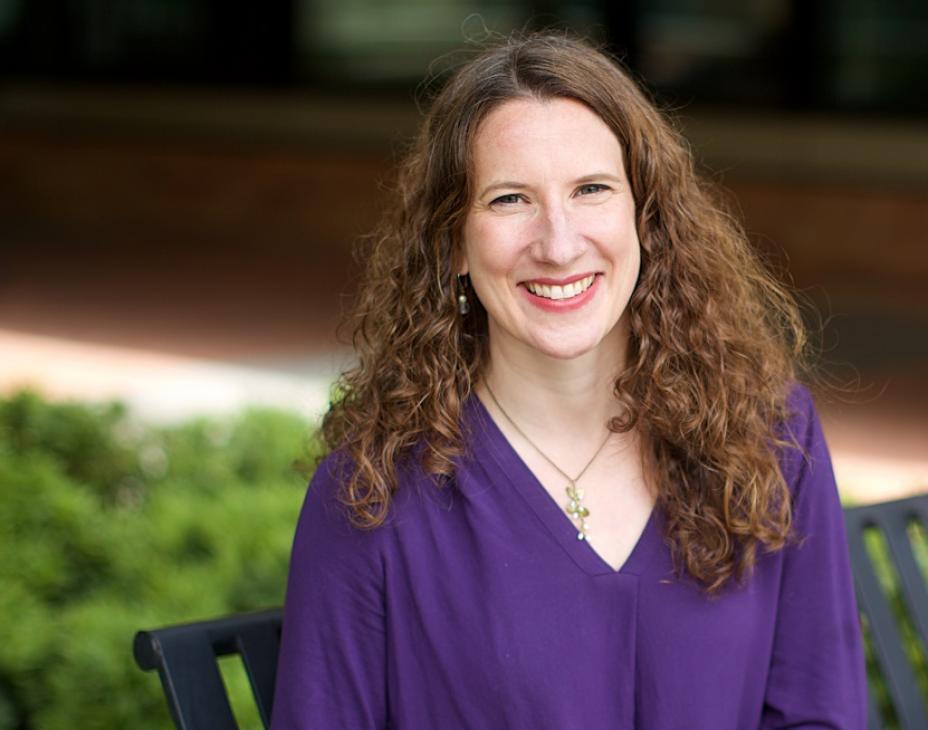 Dr. Stacey Cohen sitting outdoors on a bench in front of a green bush. She is smiling facing forward. She has shoulder-length brown hair and an indigo blouse.