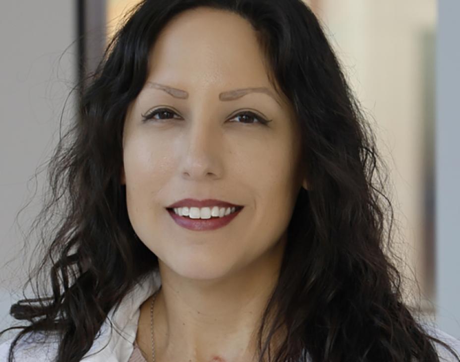 Headshot of Dr. Dionisia Quiroga. She is smiling facing forward in a clinical setting and has dark-brown hair at shoulder-length.