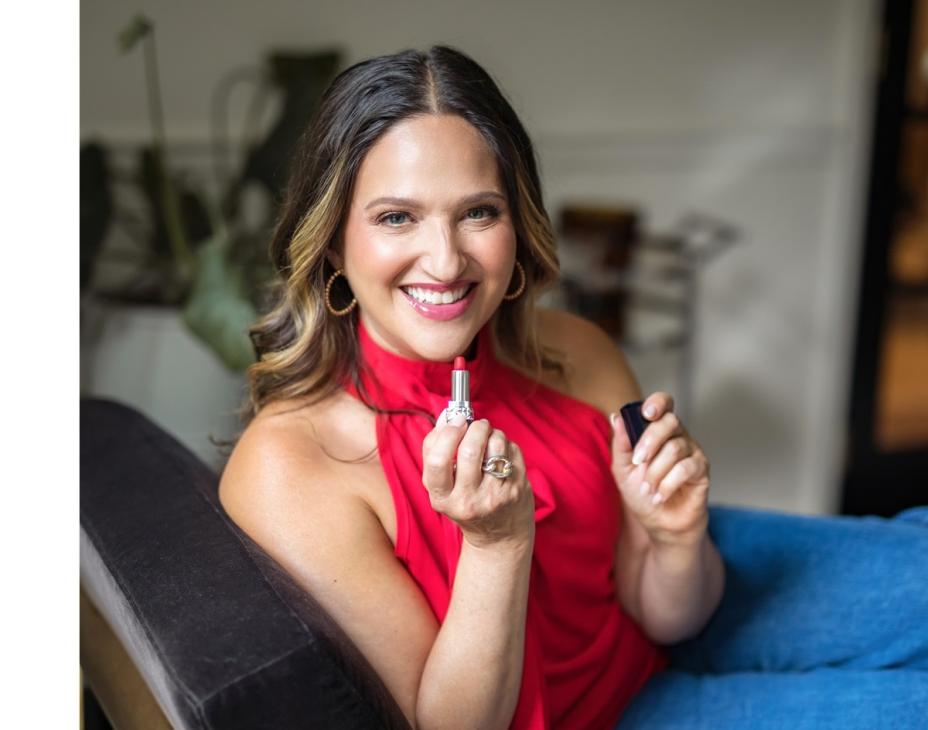 Melissa Berry holding lipstick and smiling facing forward. She's wearing a red shirt and blue jeans.