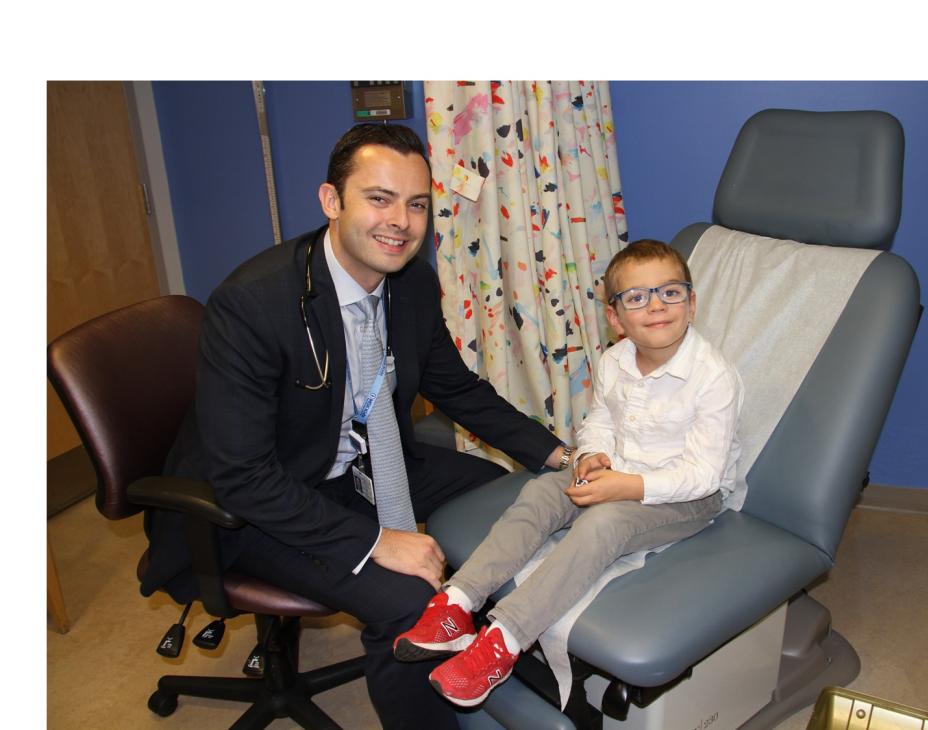 Dr. Michael Kinnaman (left) in the clinic and sitting with Calum, his patient, a 6-year-old who is sitting in the clinic chair. Both are smiling and facing forward.