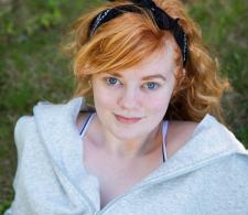 Anya Magnuson smiling, looking upwards in an outdoor, grassy environment. She has blonde hair and is wearing a white jacket.