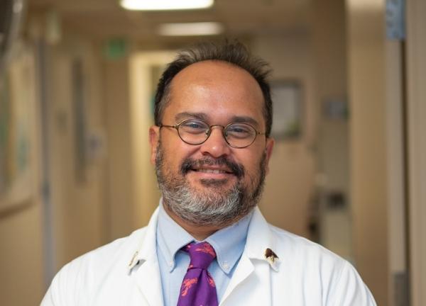 Dr. Flavio Rocha facing forward and smiling. He is wearing a white coat. He's in a well-lit hallway that appears to be in a hospital or clinic.