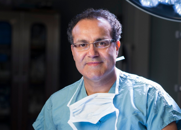 Headshot of Dr. Alfredo Quiñones-Hinojosa in surgical/medical scrubs. He's wearing glasses and has a white mask hanging on his chest, and he's standing in a surgery room.