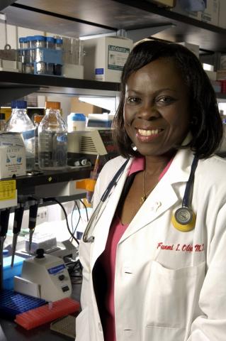 Dr. Olopade smiling at the camera, wearing a white coat while in her laboratory, with chemical equipment in the background.