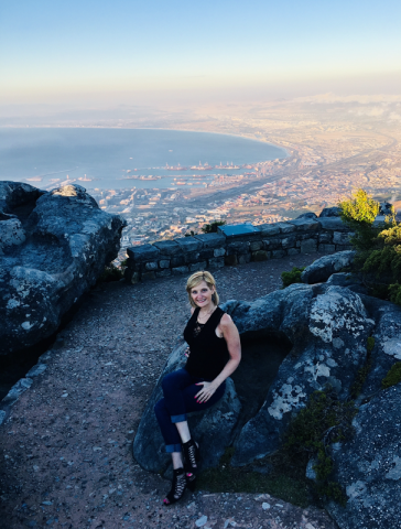 Brenda Brody in athletic gear sitting atop a path overlooking a scenic, atmospheric view of Cape Town, supplemented with a soft, light-blue, cream-yellow horizon in the distance.