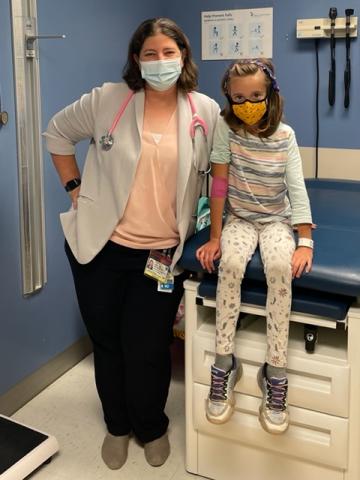 Left to right: Dr. Jessica Lake and her patient, Mia. Both are in a clinical setting, wearing masks, and smiling facing forward. Dr. Lake is standing next to Mia, who is sitting on a patient bench.