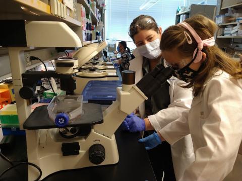 Dr. Jessica lake with her patient, Mia, in a research lab. Mia is looking in a microscope while Dr. Lake mentors and observes.