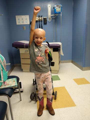 Mia holding her arm up like superwoman. She is in a clinical setting, smiling in a celebratory way.