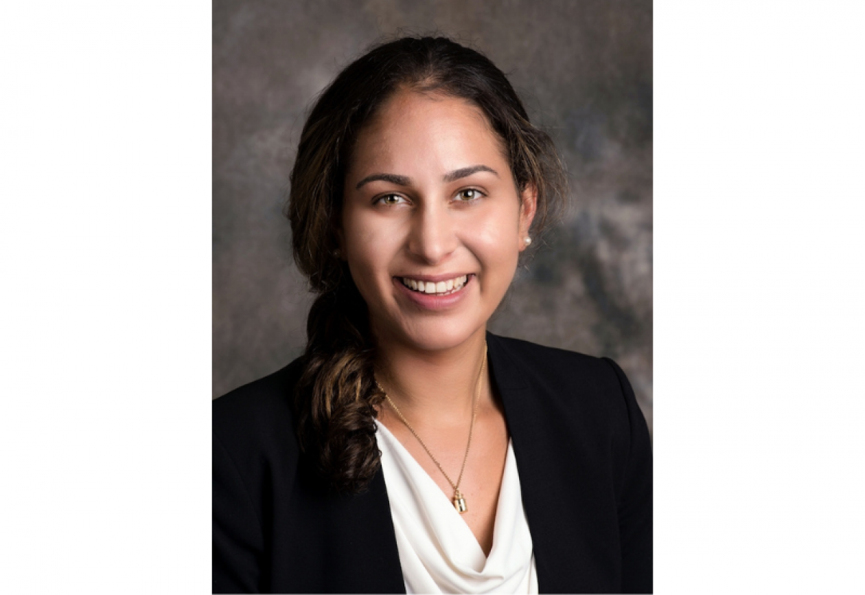 Headshot of Dr. Eleanor Taranto. She is smiling facing forward against a gray background and has shoulder-length brown hair, and is wearing a black blazer over a white blouse with a necklace.