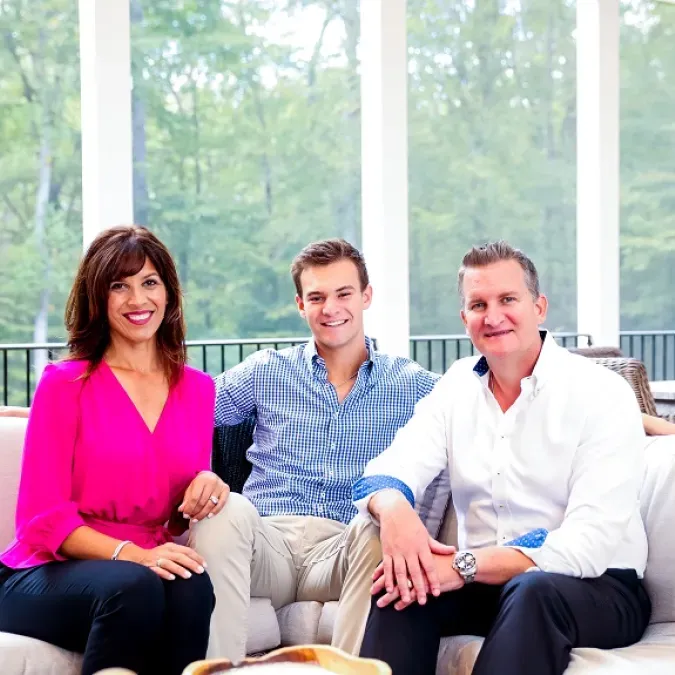 Left to right: Nancy Berry, Jake Berry, Alex Berry. All three are seated facing forward and smiling, located in a screened-in sun-room with white columns and green trees visible in the background. Nancy is wearing a bright pink shirt with dark blue pants. Jake is wearing a light blue shirt with khaki plants. Alex is wearing a white shirt with black pants.