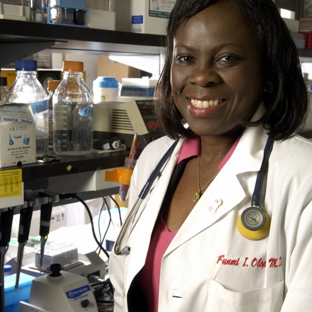 Dr. Olopade smiling at the camera, wearing a white coat while in her laboratory, with chemical equipment in the background.