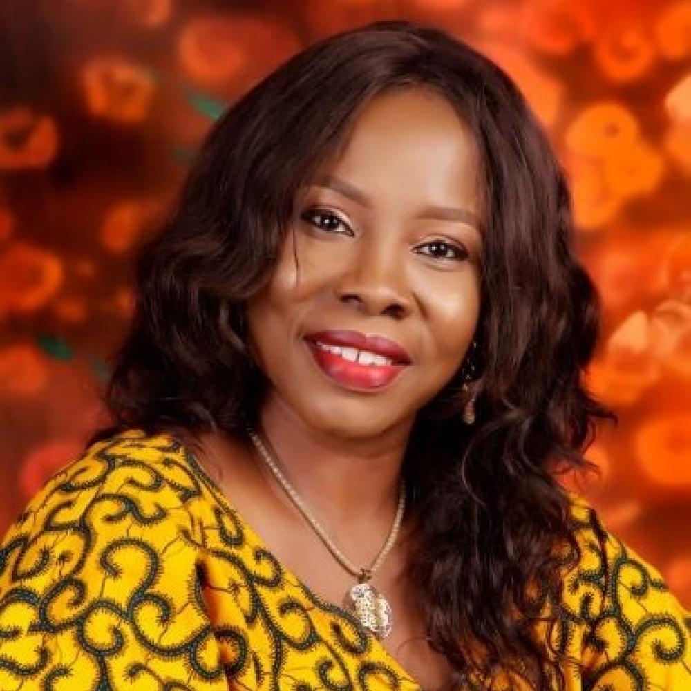 Headshot of Dr. Tonia Onyeka smiling. Background is bright, deep orange. Dr. Onyeka is wearing a gold-colored long-sleeve shirt with an intricate pattern colored dark brown to black.