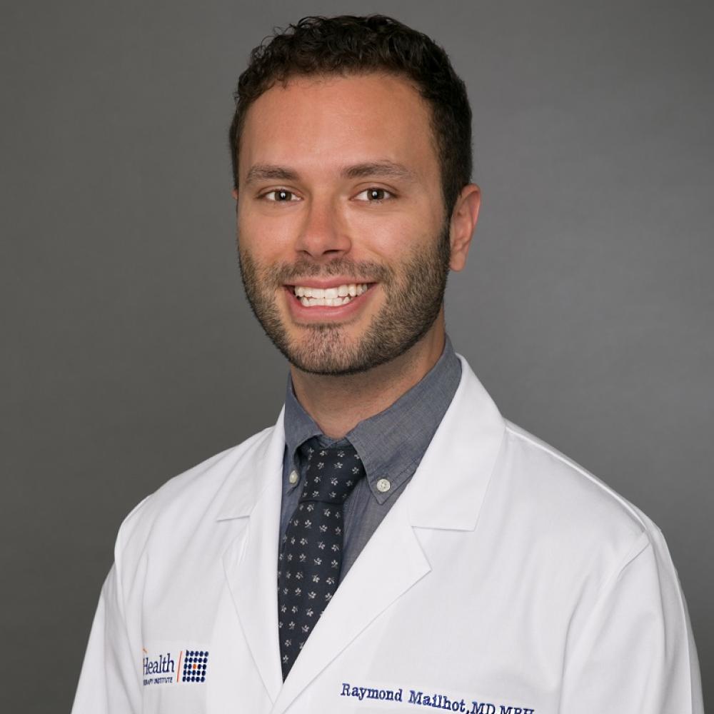 Headshot of Dr. Raymond Mailhot. He is wearing a white coat with his name engraved on it, as well as a dark gray shirt and tie. His hair is dark brown to black and he is smiling facing forward.