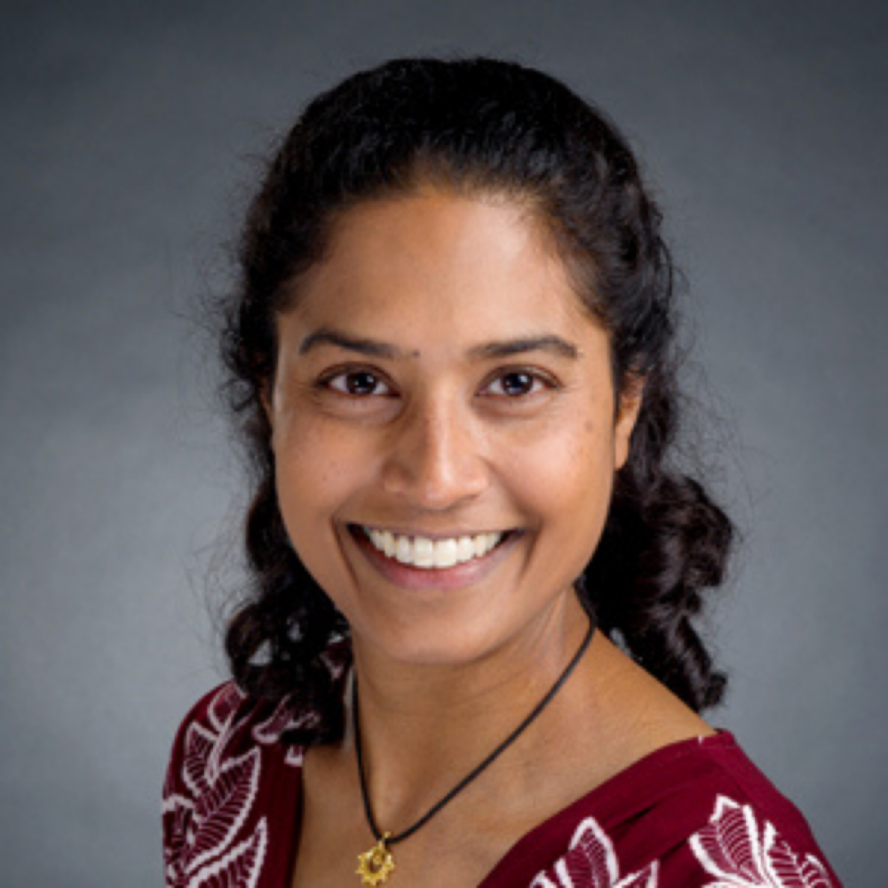 Dr. Sheena Mukkada facing forward and smiling. She has shoulder-length black hair and is wearing a dark-red shirt with a black necklace, against a plain gray background.