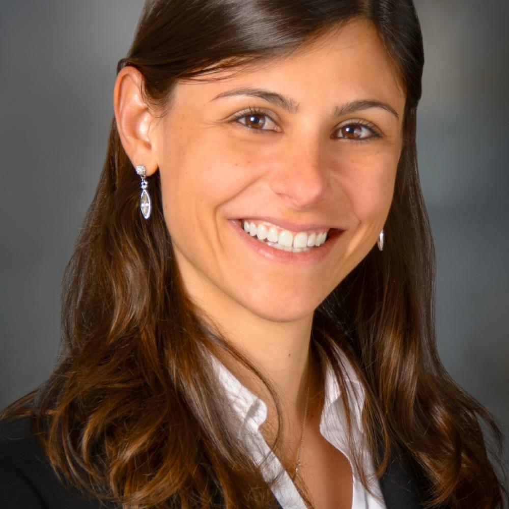 Dr. Ferrarotto headshot. She is wearing a black blazer and has shoulder-length brown hair. She is smiling facing forward against a gray background.