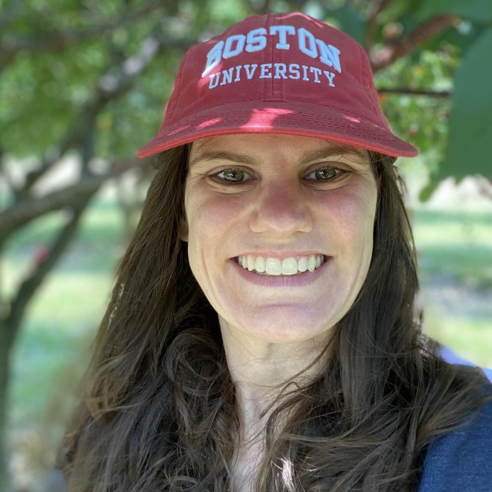Carly Flumer wearing a red Boston University hat, smiling facing forward while taking a selfie outside in a green environment. She has long brown hair and is wearing a blue shirt.
