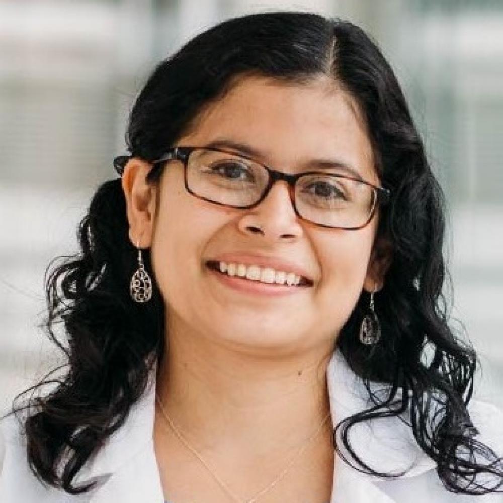 Headshot of Dr. Jenny Ruiz. She has shoulder-length black hair and is wearing glasses, in a white coat. She is smiling facing forward in a bright hallway.