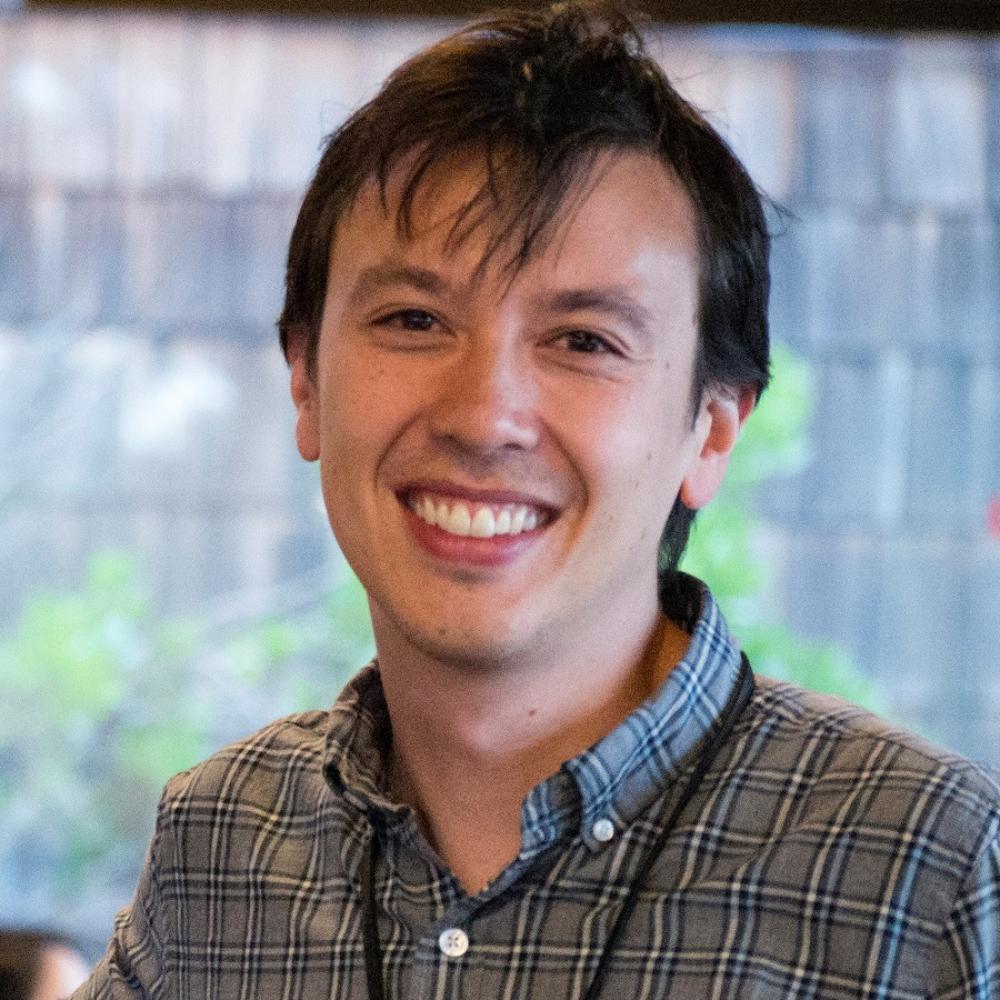 Dr. David Kurtz wearing a dark green shirt and lanyard in a brightly-lit indoor environment. He has short dark-brown hair and is facing forward.