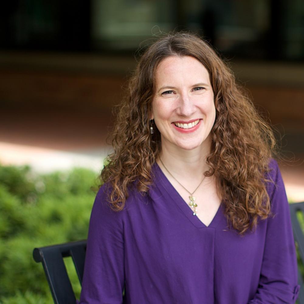Dr. Stacey Cohen sitting outdoors on a bench in front of a green bush. She is smiling facing forward. She has shoulder-length brown hair and an indigo blouse.