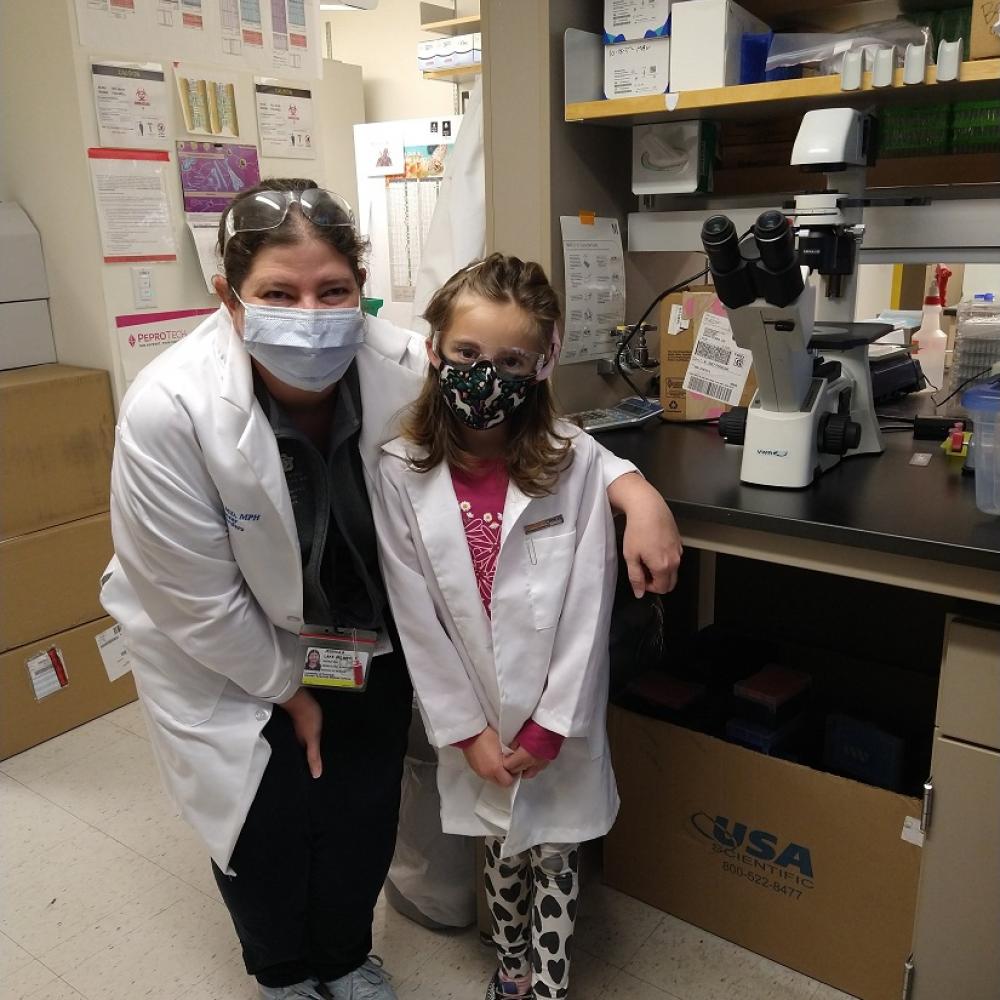 Right to left: Dr. Jessica Lake and her patient, Mia. Both are wearing masks and doctor white coats, smiling facing forward.