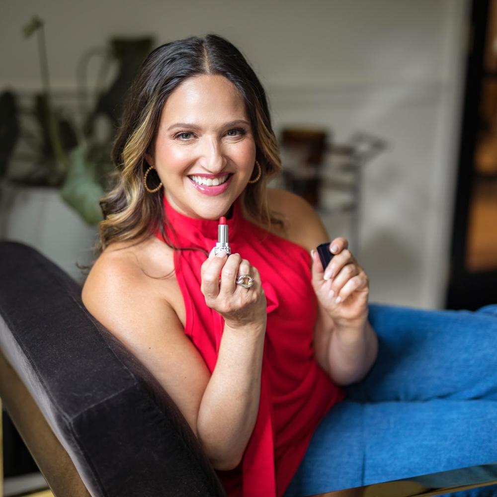 Melissa Berry holding lipstick and smiling facing forward. She's wearing a red shirt and blue jeans.