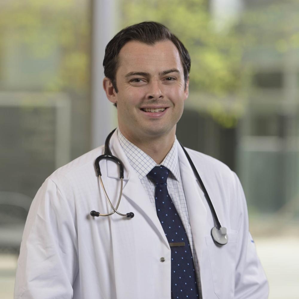 Dr. Michael Kinnaman in a doctor's coat with a stethoscope, smiling facing forward in an outdoor setting