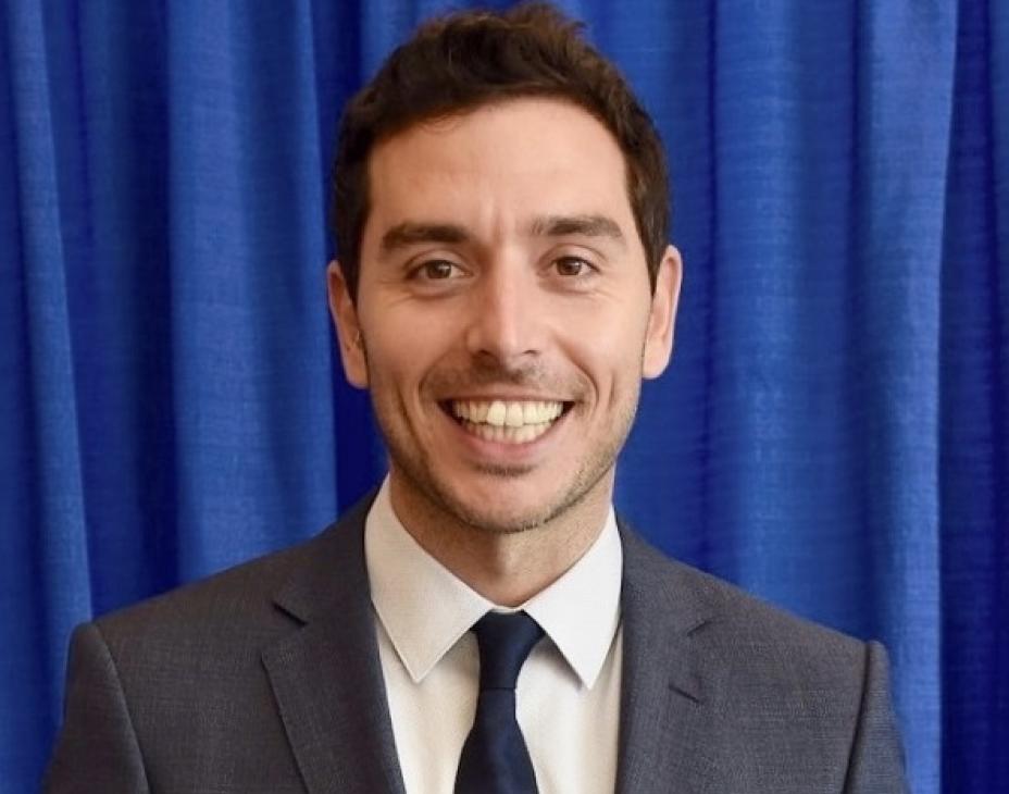 Headshot of Dr. Marc Oliva smiling facing forward. He is wearing a dark-gray suit and has short dark-brown hair. He is in front of a blue curtain background.