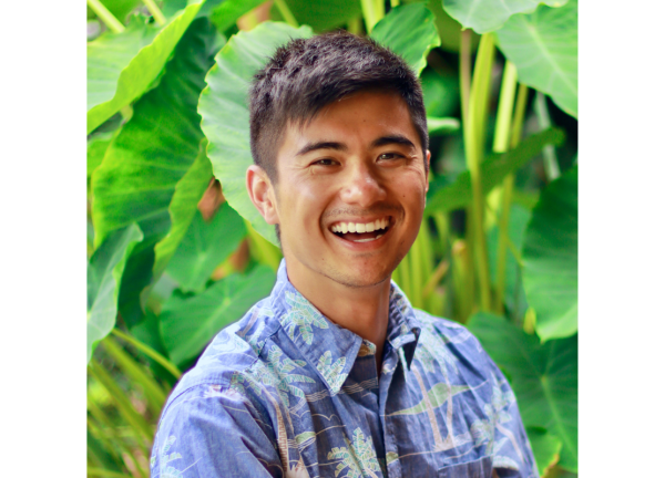 Dr. Kekoa Taparra headshot. He is smiling facing, wearing a light-blue shirt, forward against a background of bright-green tropical leaves. 
