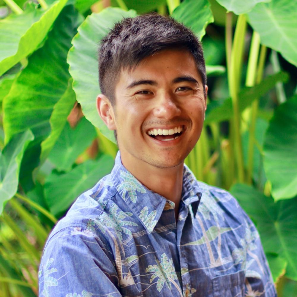Dr. Kekoa Taparra headshot. He is smiling facing, wearing a light-blue shirt, forward against a background of bright-green tropical leaves. 