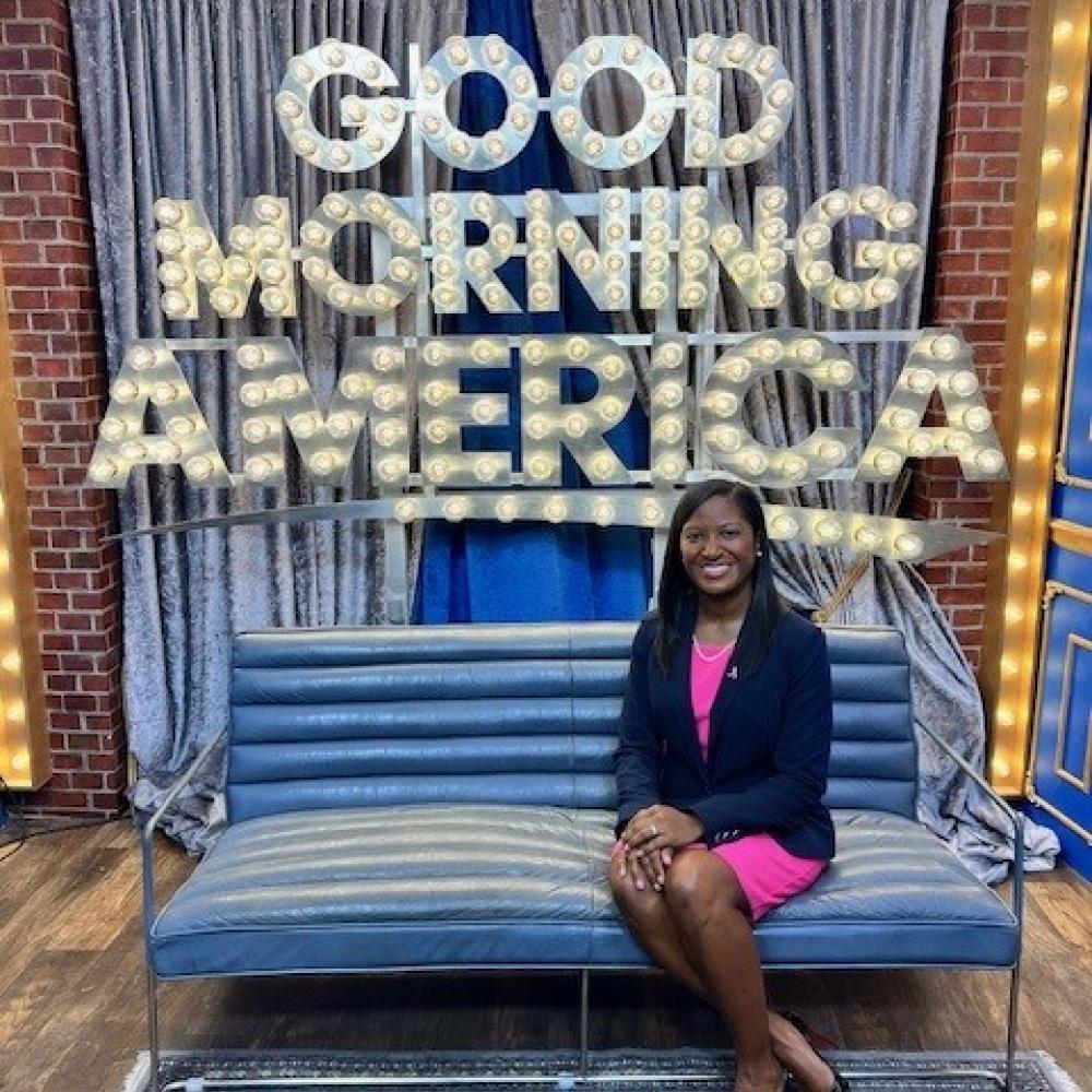 Dr. Erica Stringer-Reasor smiling facing forward against a 'Good Morning America' sign. She has shoulder-length black hair.